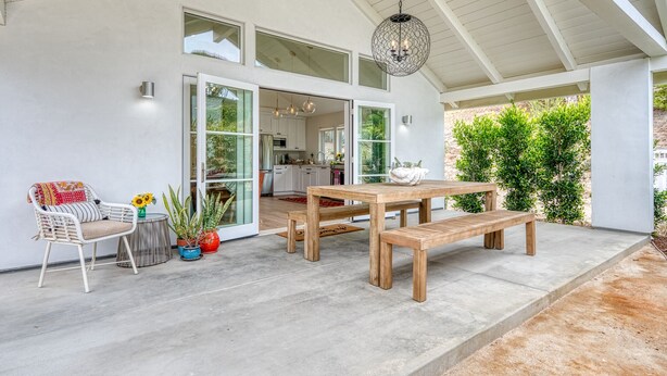 Un porche cubierto con una mesa y bancos de madera, una silla blanca y plantas en macetas.