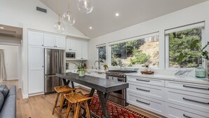 Una cocina moderna con armarios blancos, electrodomésticos de acero inoxidable, una isla oscura y un amplio ventanal con vistas a la vegetación.