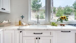 Una cocina con armarios blancos, encimera de mármol, lavabo y ventana con vistas a árboles y una valla.