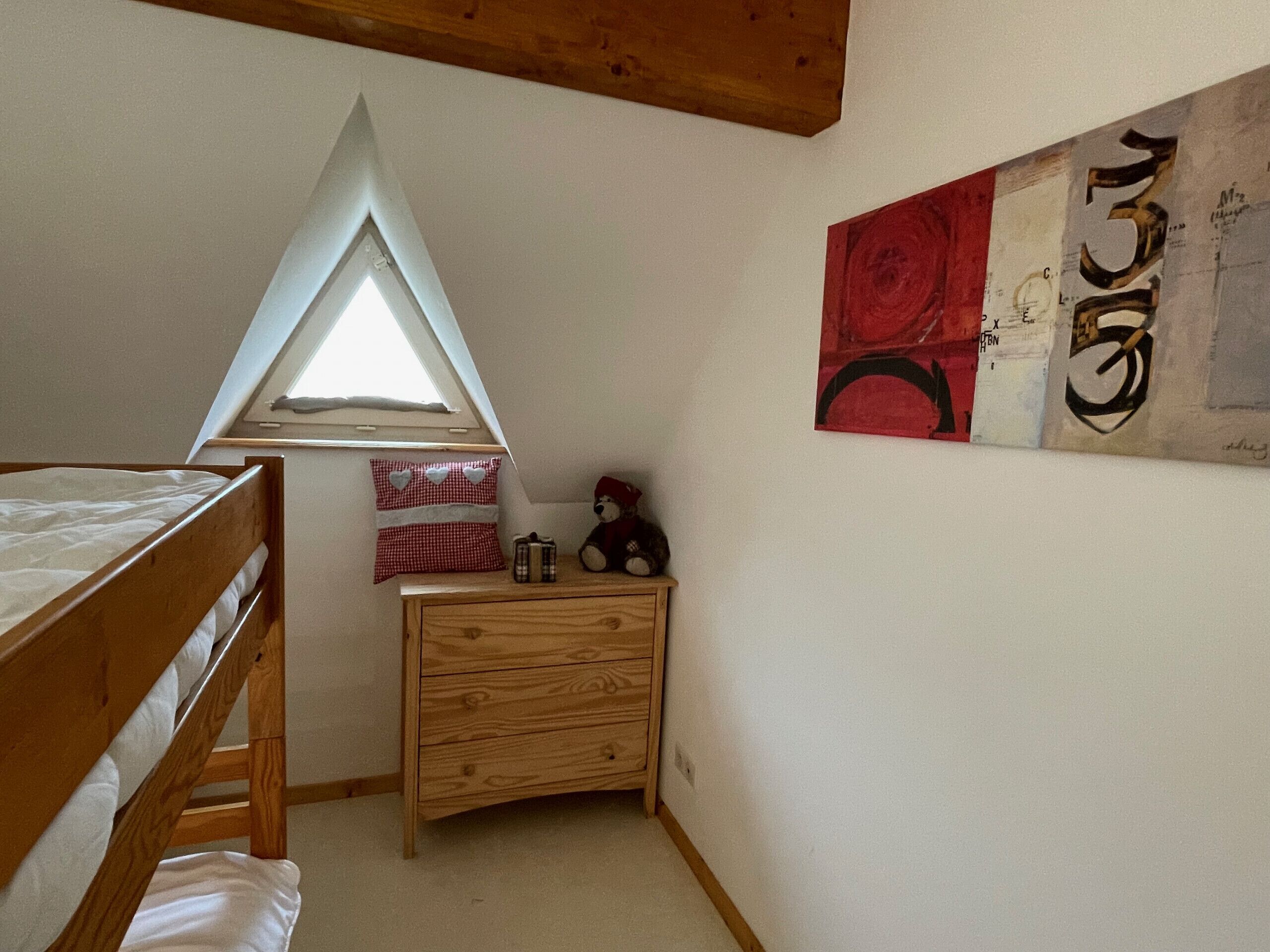A bedroom with a bunk bed, a small wooden dresser, a window, and a wall with abstract art.