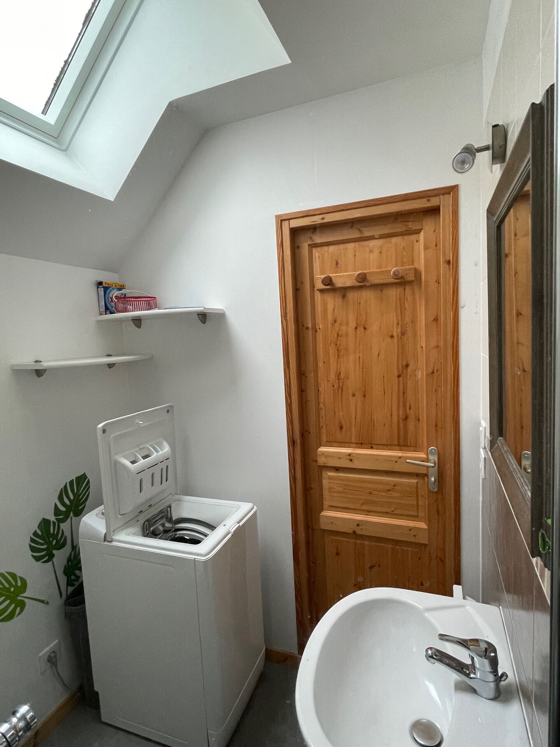 Une petite salle de bain dotée d’un puits de lumière, d’un lave-linge blanc, d’une porte en bois et d’un lavabo avec un miroir.