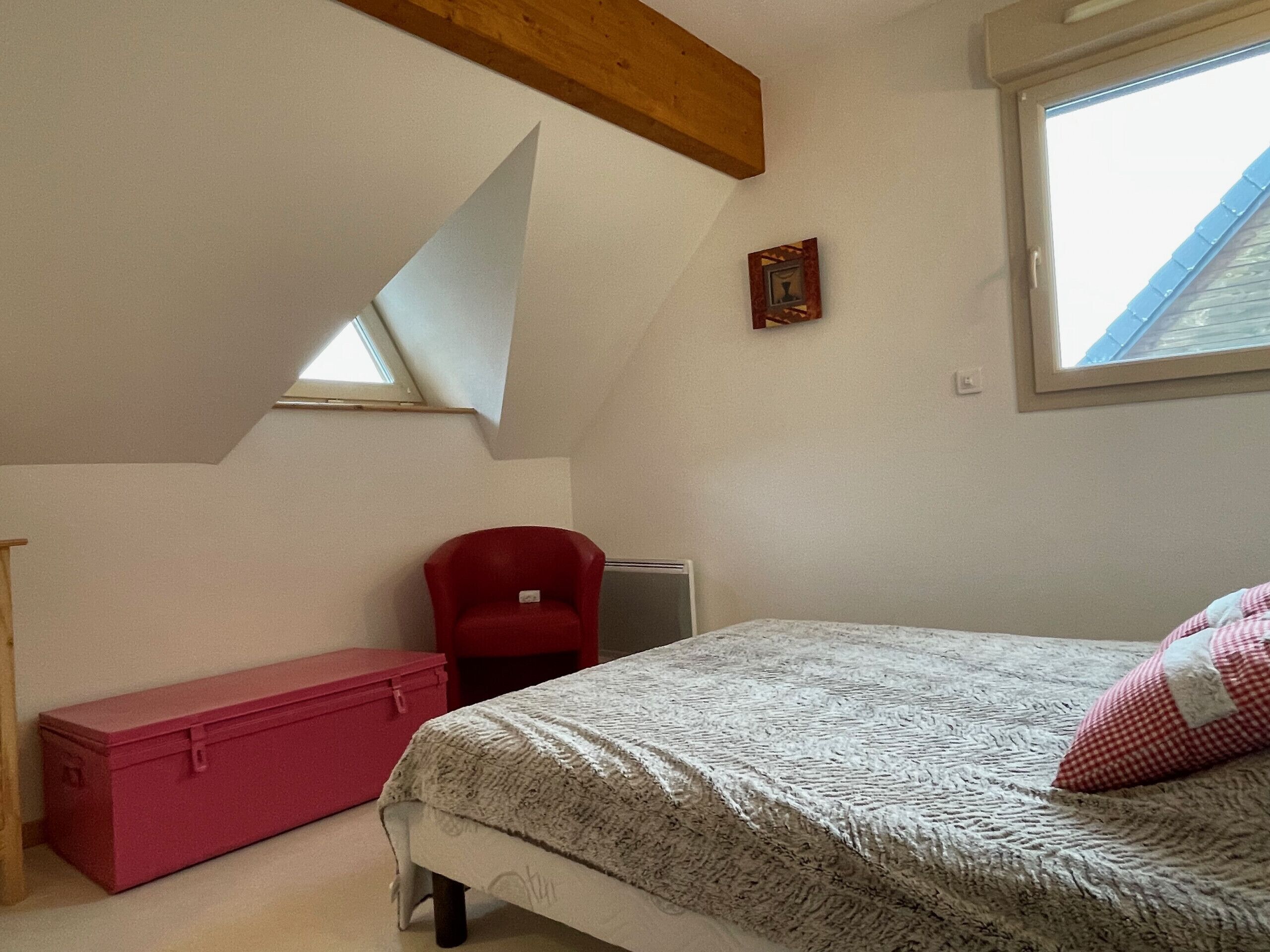 Une chambre avec un plafond en pente, une chaise rouge, un lit recouvert d’une couverture à motifs, une boîte de rangement rose et un petit tableau encadré accroché au mur.