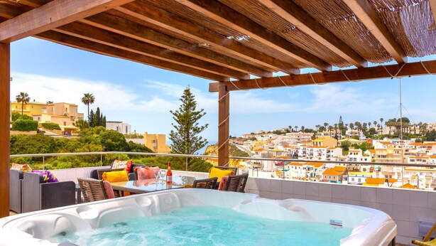 Un jacuzzi con una pérgola de madera encima, rodeado de mobiliario exterior y vistas a un pueblo costero.
