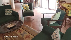 Ein geräumiges Wohnzimmer mit hoher Decke, grünen Sofas, einem Couchtisch und Blick durch eine Glastür in einen Außenbereich.