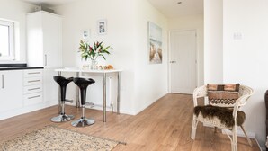 Una cocina moderna con armarios blancos, una mesa de comedor, dos taburetes negros, una silla blanca con un cojín estampado y suelo de madera.
