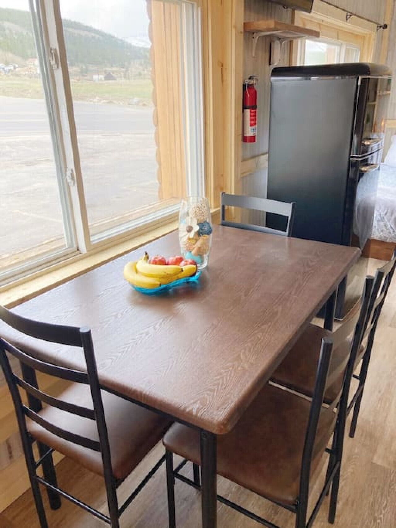 A wooden dining table with chairs, a window with a view of a parking lot and hills, and a refrigerator in the background.