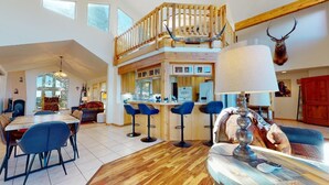 Een ruime woonkamer met een hoge plafond, een eettafel met stoelen, een bar met krukken, en een opgezette hertenkop aan de muur.