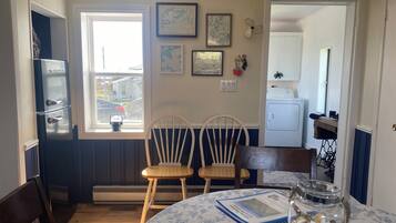 A dining area with two wooden chairs, a round table with a floral tablecloth, a glass jar with seashells, and a refrigerator in the background.