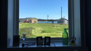 A window with a view of a grassy area, a lake, and houses in the distance. - Seaside Cottage in Beautiful Charlo’s Cove (Charlo's Cove)