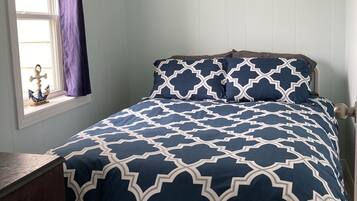 Un dormitorio con cama, una ventana con cortinas, un adorno en la pared y una pequeña mesa con un objeto decorativo.