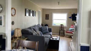 A cozy living room with a grey sofa, a wooden chair, a small table with a basket, and a window with a view of the outdoors. - Seaside Cottage in Beautiful Charlo’s Cove (Charlo's Cove)