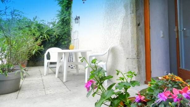 Um pátio externo com mesa e cadeiras brancas, rodeado por vasos com plantas e flores.