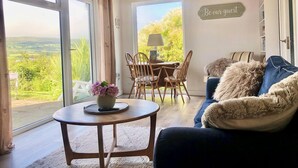 A cozy living room with a view of a lush landscape, a dining area with a table and chairs, and a sign that reads "Be our guest". - 1 Bed Flat - Sleeps 5 - Parking - Garden - Pets (Larne)