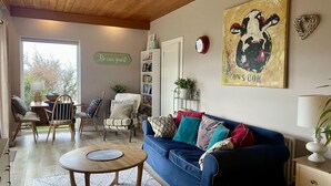 A cozy living room with a blue sofa, wooden chairs, a round coffee table, and a bookshelf. A painting of a cow is on the wall, and there's a 'Be our guest' sign. - 1 Bed Flat - Sleeps 5 - Parking - Garden - Pets (Larne)