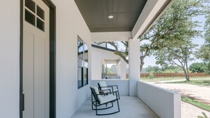 Un espace extérieur abrité, doté d’un mur blanc, d’une porte et d’une fenêtre. On y trouve une chaise avec un coussin et une petite table.