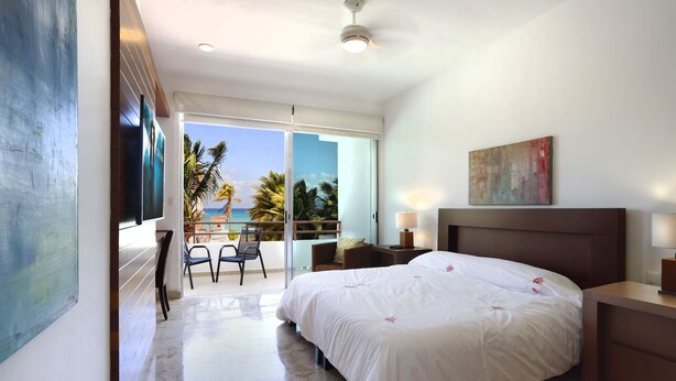 Ein Hotelzimmer mit einem großen Bett, einem Schreibtisch, einem Sessel, einem Fernseher und einem Balkon mit Blick auf Palmen und das Meer.