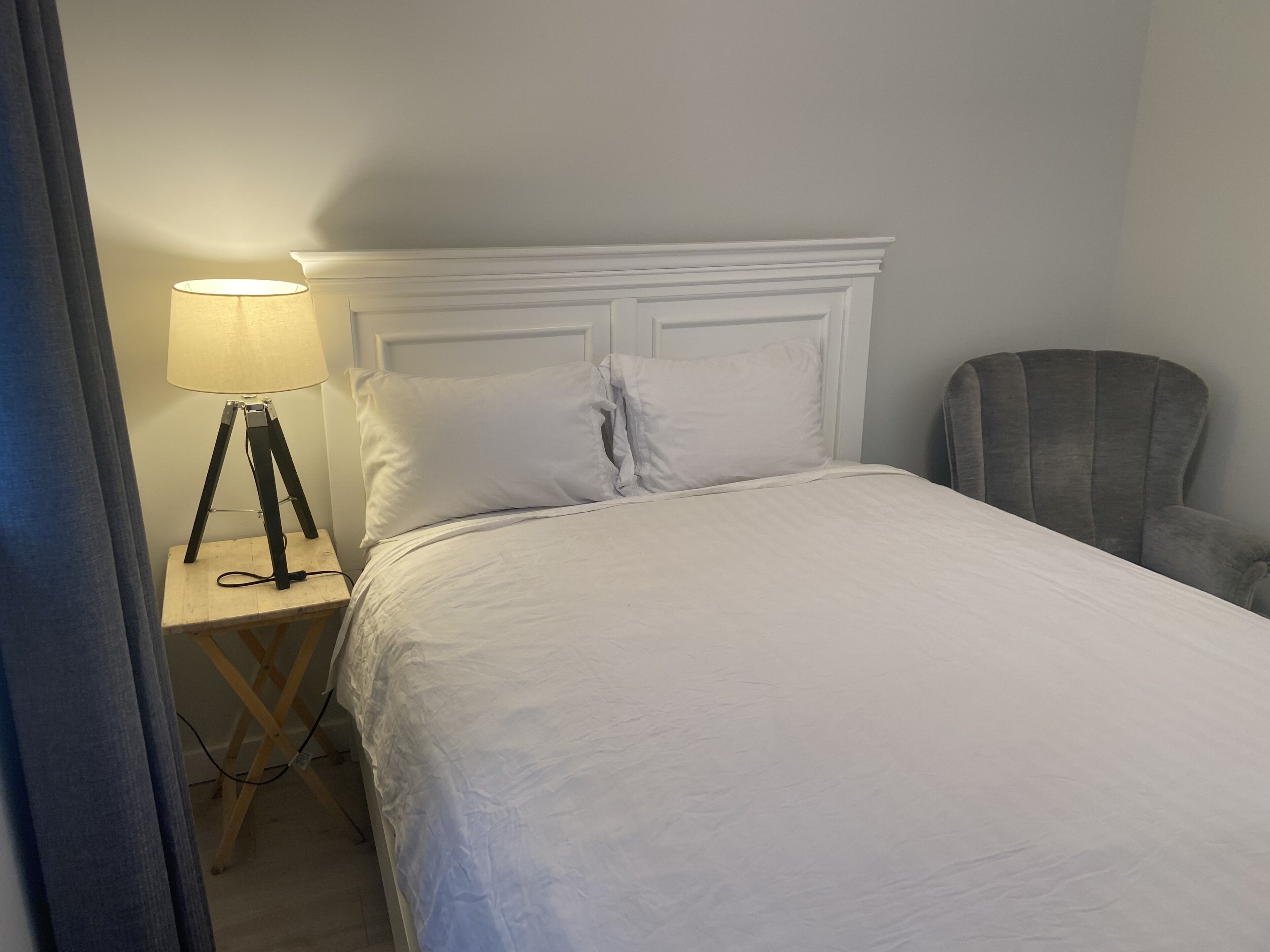 Un lit bien fait, avec une literie blanche, une lampe sur une table de chevet en bois et un fauteuil gris.