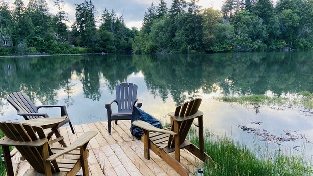 Eine Holzterrasse mit Adirondack-Stühlen und einem Tisch, mit Blick auf einen stillen See, umgeben von dichtem Wald.