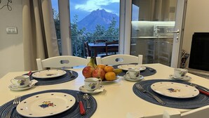 Een eettafel gedekt met borden, bestek en een fruitschaal, met uitzicht op een berg vanuit het raam.