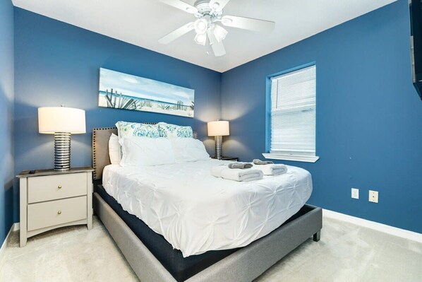 A bedroom with a bed, two bedside tables with lamps, a ceiling fan, a window with blinds, and a beach-themed painting on the wall.