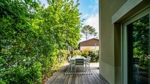 Une terrasse en bois avec une table et des chaises, entourée d’une végétation luxuriante et d’un bâtiment doté d’une grande fenêtre.