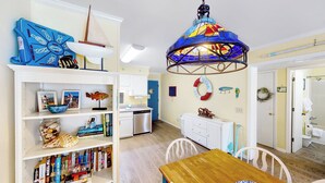 A room with a wooden dining table, white chairs, a bookshelf with books and decorative items, a sailboat model, a colorful hanging light fixture, and a blue wall with nautical-themed decorations.