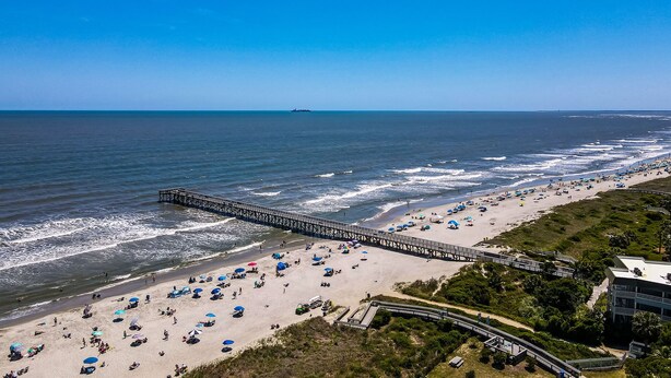 A beach with a long pier extending into the ocean, sandy beach with umbrellas, and a clear blue sky.