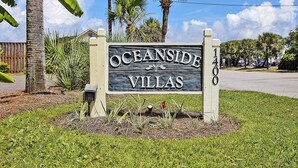 A sign for 'Oceanside Villas' with a wooden texture, surrounded by grass and palm trees.
