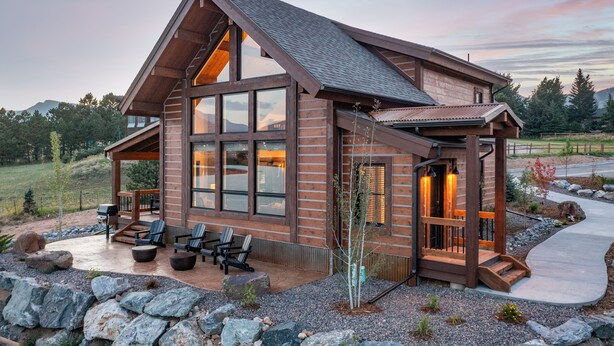 A modern log cabin with large windows, a covered patio with outdoor seating, and a stone pathway leading to the entrance.