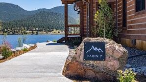 A cabin labeled 'CABIN 7' with a mountain view, wooden exterior, and a concrete pathway leading to the entrance.