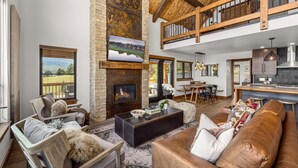 A spacious living room with a fireplace, a large couch, and a dining area with a high ceiling and exposed beams.