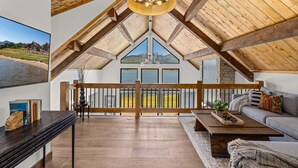 A spacious loft with a high ceiling, wooden beams, a large window overlooking a scenic landscape, a cozy seating area with a sofa and coffee table, and a wooden railing along the balcony.