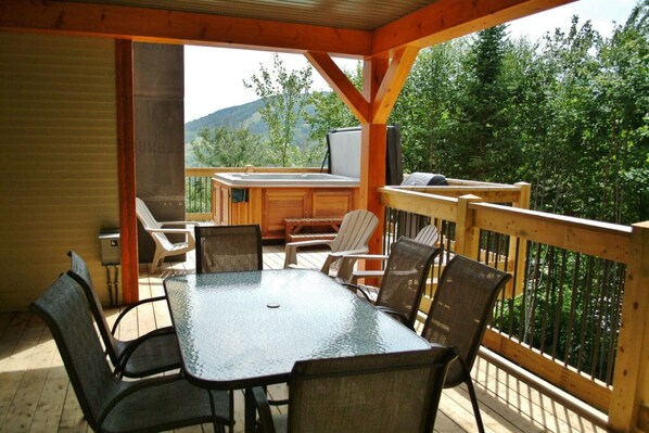 A covered outdoor deck with a table and chairs, a hot tub, and a view of the surrounding forest. - 14- Cottage for rent in Stoneham (CITQ: 245486) (Stoneham-et-Tewkesbury)