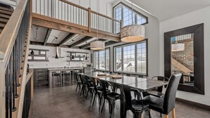 A spacious dining area with a long table and black chairs, a kitchen with white cabinets, and large windows offering a view of the outdoors. - 14- Cottage for rent in Stoneham  (CITQ: 245486) (Stoneham-et-Tewkesbury)