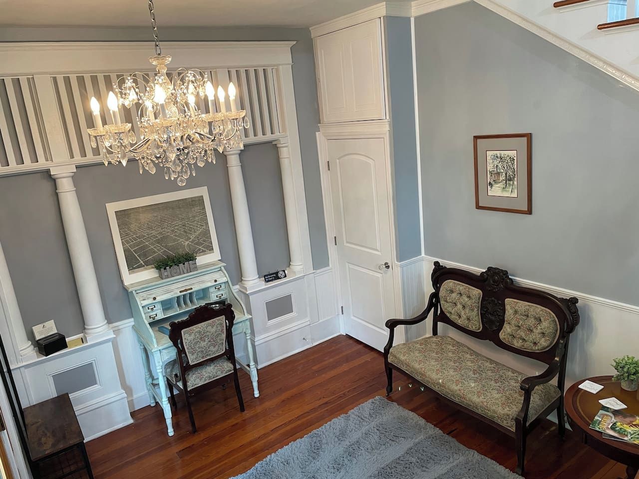 A room with a wooden floor, a white console table, a dark wooden bench, a chandelier, and a framed picture on the wall.