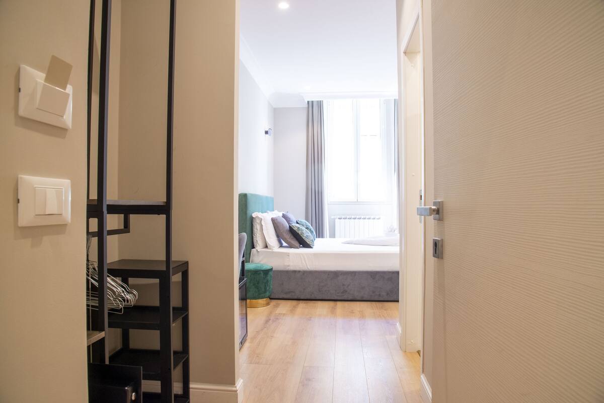 A narrow hallway leading to a bedroom with a bed, a nightstand, and a window with curtains. There is a black shelving unit with clothes on it.