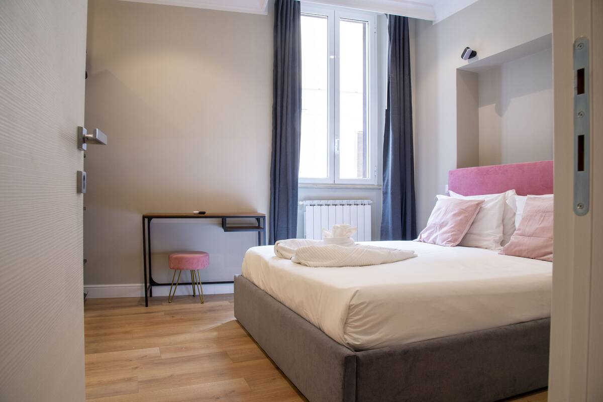 A modern bedroom with a large bed, a small desk, and a window with curtains.