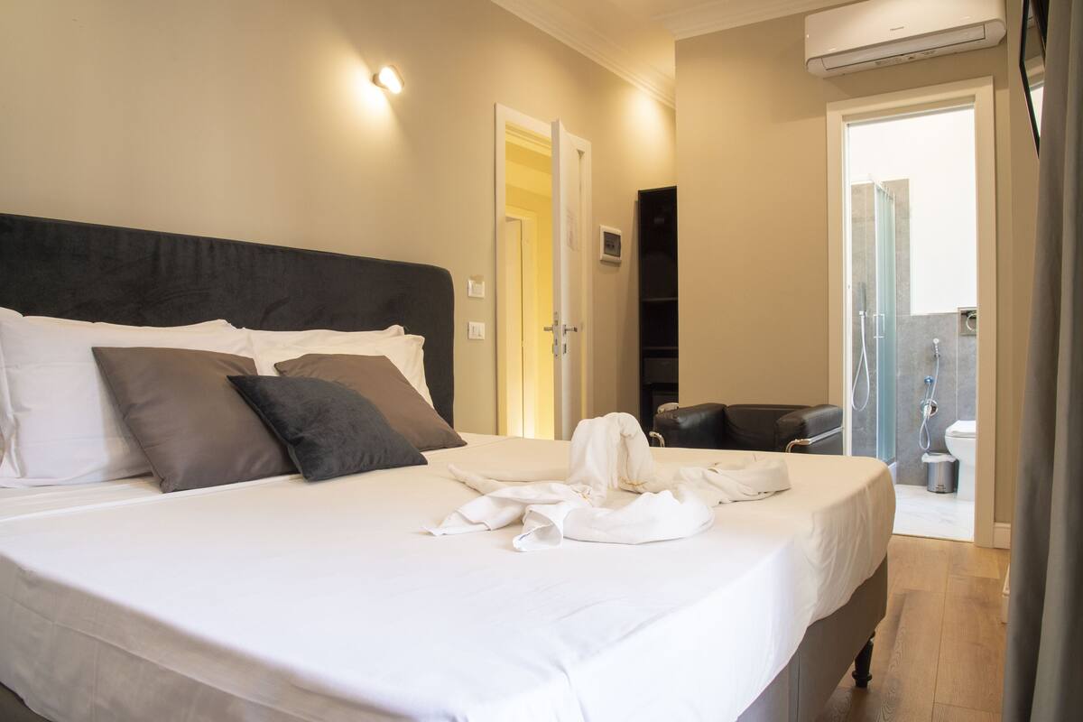 A hotel room with a large bed, a black headboard, and a shower area visible through an open door.