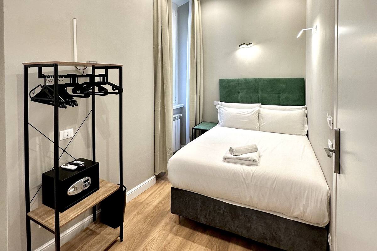 A hotel room with a bed, a coat rack with a radio, a small table, and a chair.