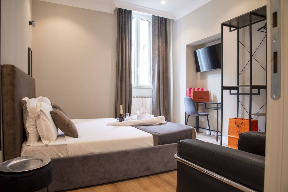 A modern hotel room with a bed, a seating area, a television, and a wardrobe.