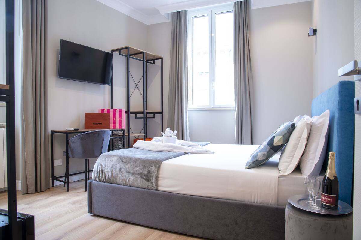 A modern hotel room with a bed, a desk, a TV, and a bottle of champagne on a side table.
