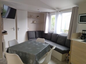 A compact living space with a dining table, a built-in bench, a flat-screen TV, a coffee maker, and a window with curtains.