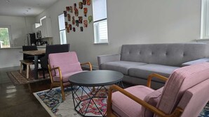 Ein Wohnzimmer mit grauem Sofa, zwei rosafarbenen Sesseln, einem kleinen runden Tisch und einem Holztisch. Im Hintergrund ist eine Küche mit weißen Schränken und einem Fenster zu sehen.