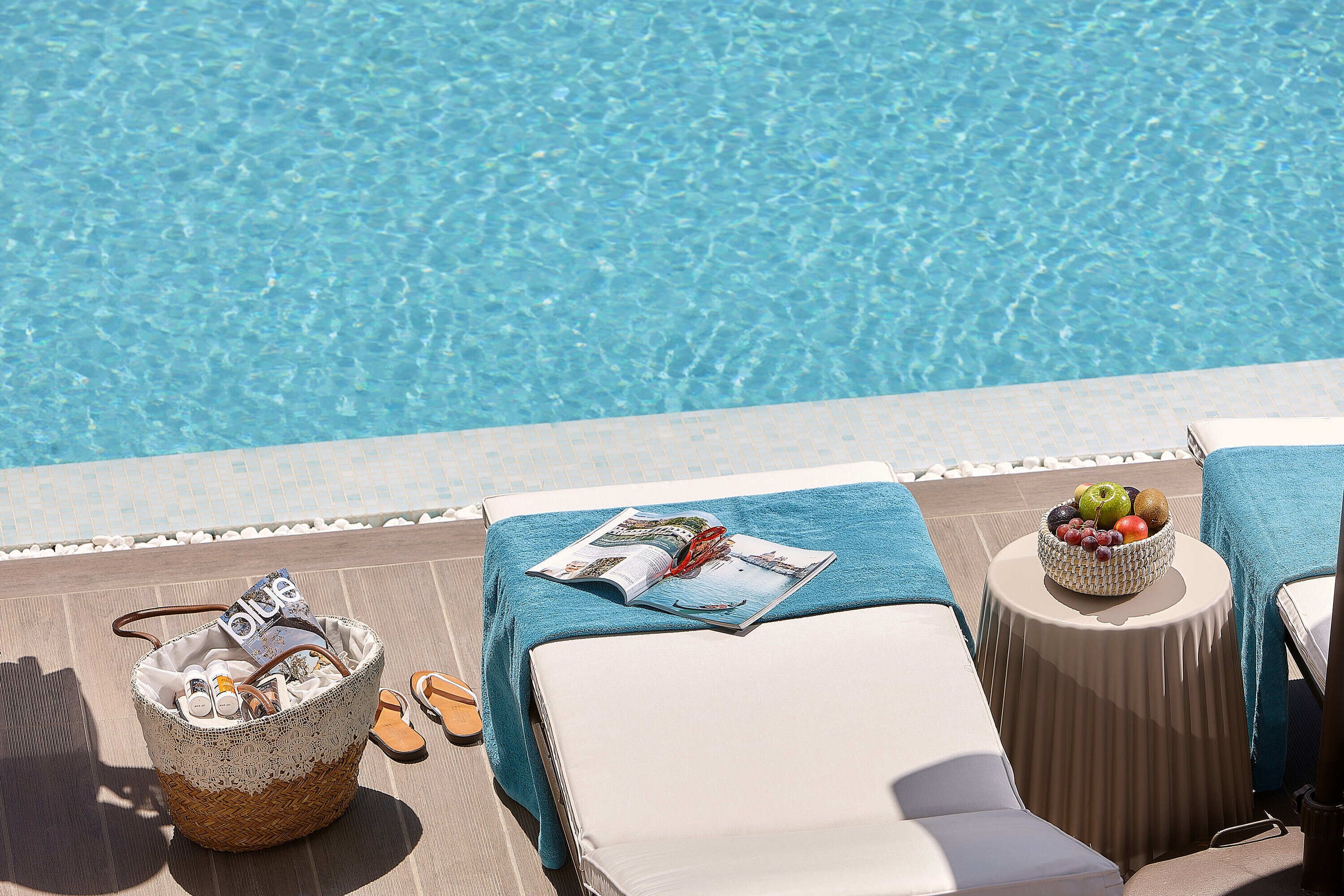A poolside area with a wicker basket containing bottles, a magazine, and flip-flops, a round table with a fruit bowl, and lounge chairs with a blue towel.