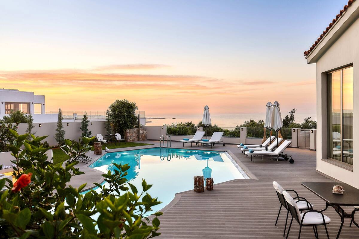 Zona de piscina en la azotea con sillones de descanso y sombrillas, rodeada de vegetación y con vistas al mar.