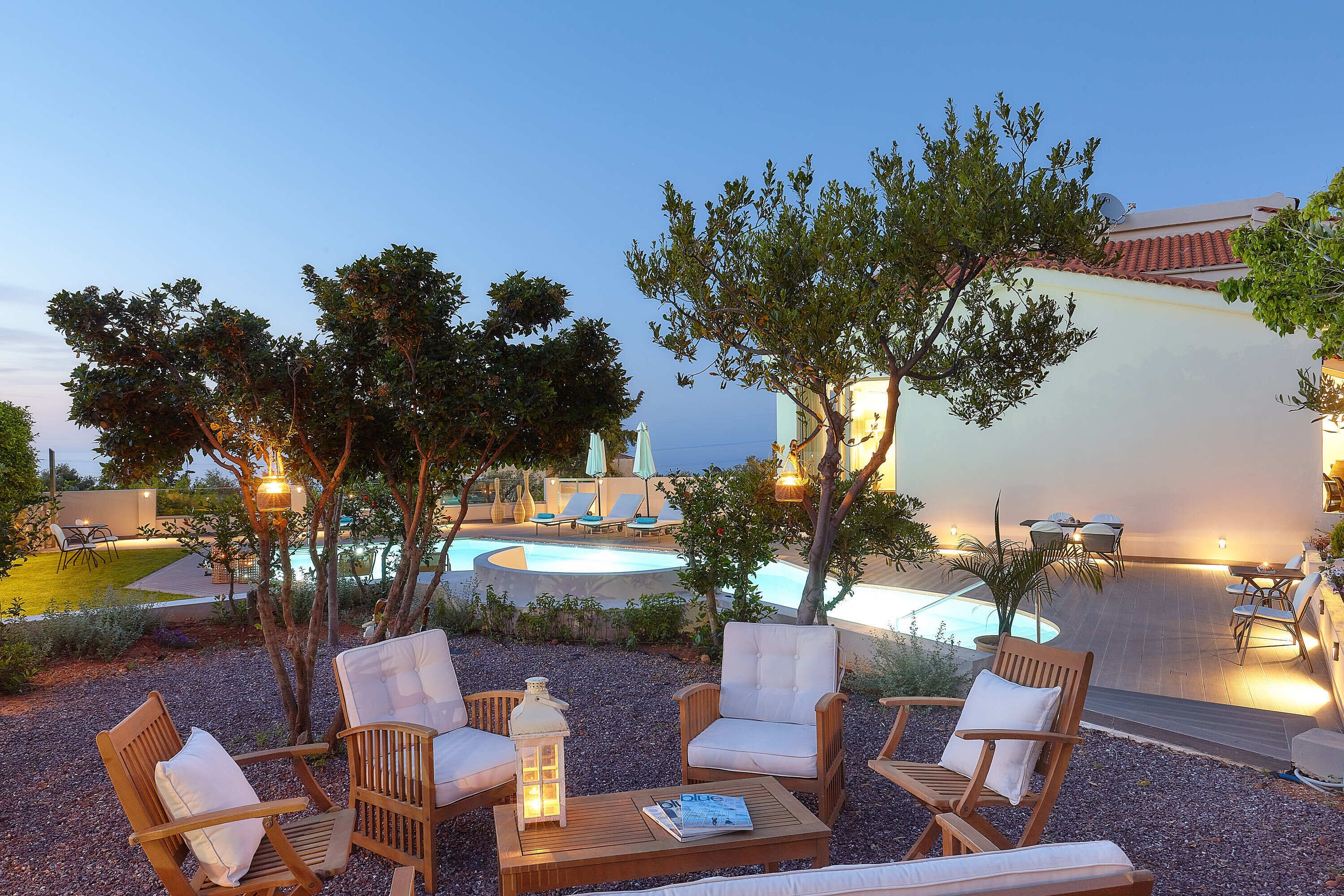 An outdoor seating area with wicker chairs and a wooden table, a pool in the background, and trees providing shade.