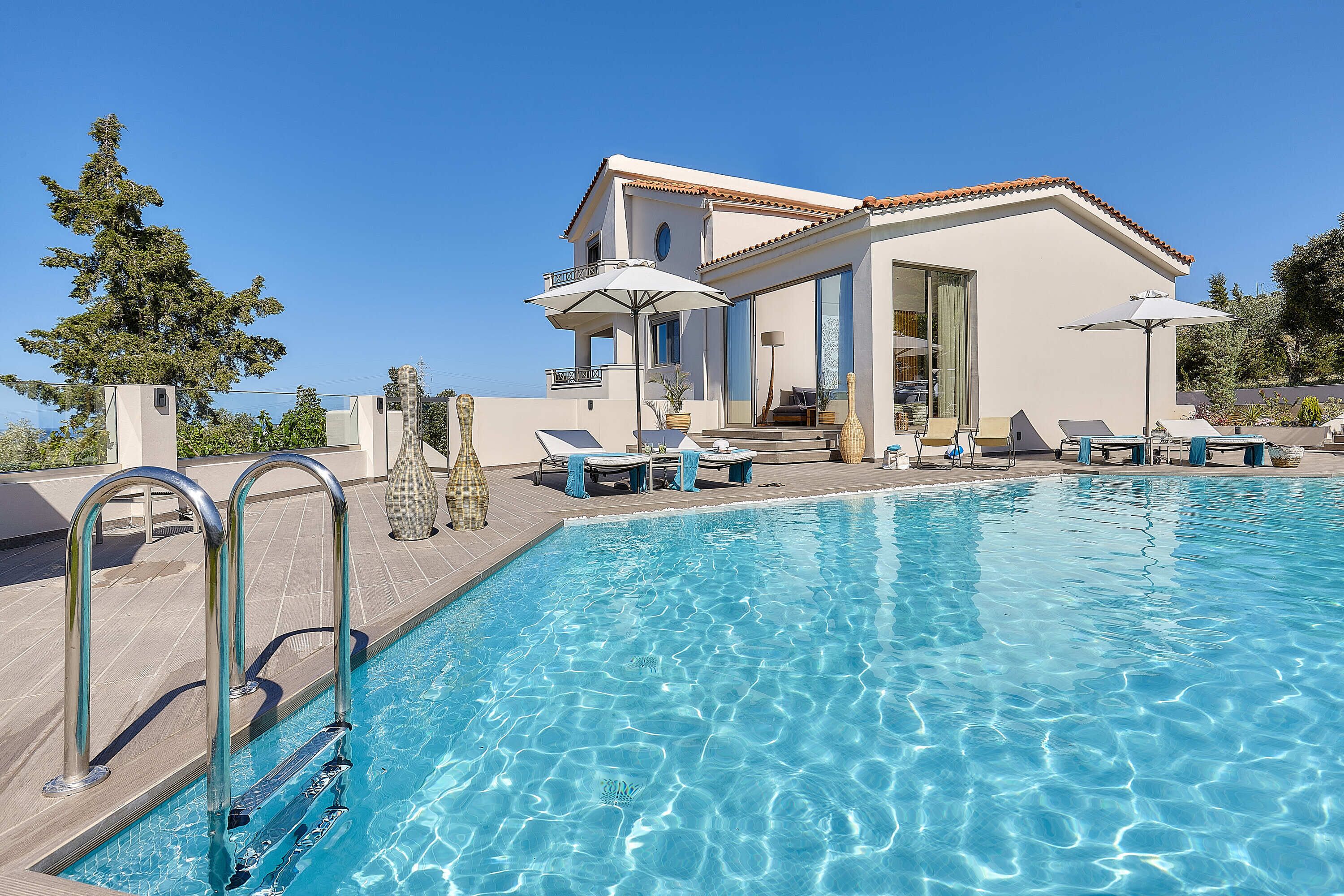 A swimming pool with lounge chairs and umbrellas, a building with a balcony, and a clear blue sky.