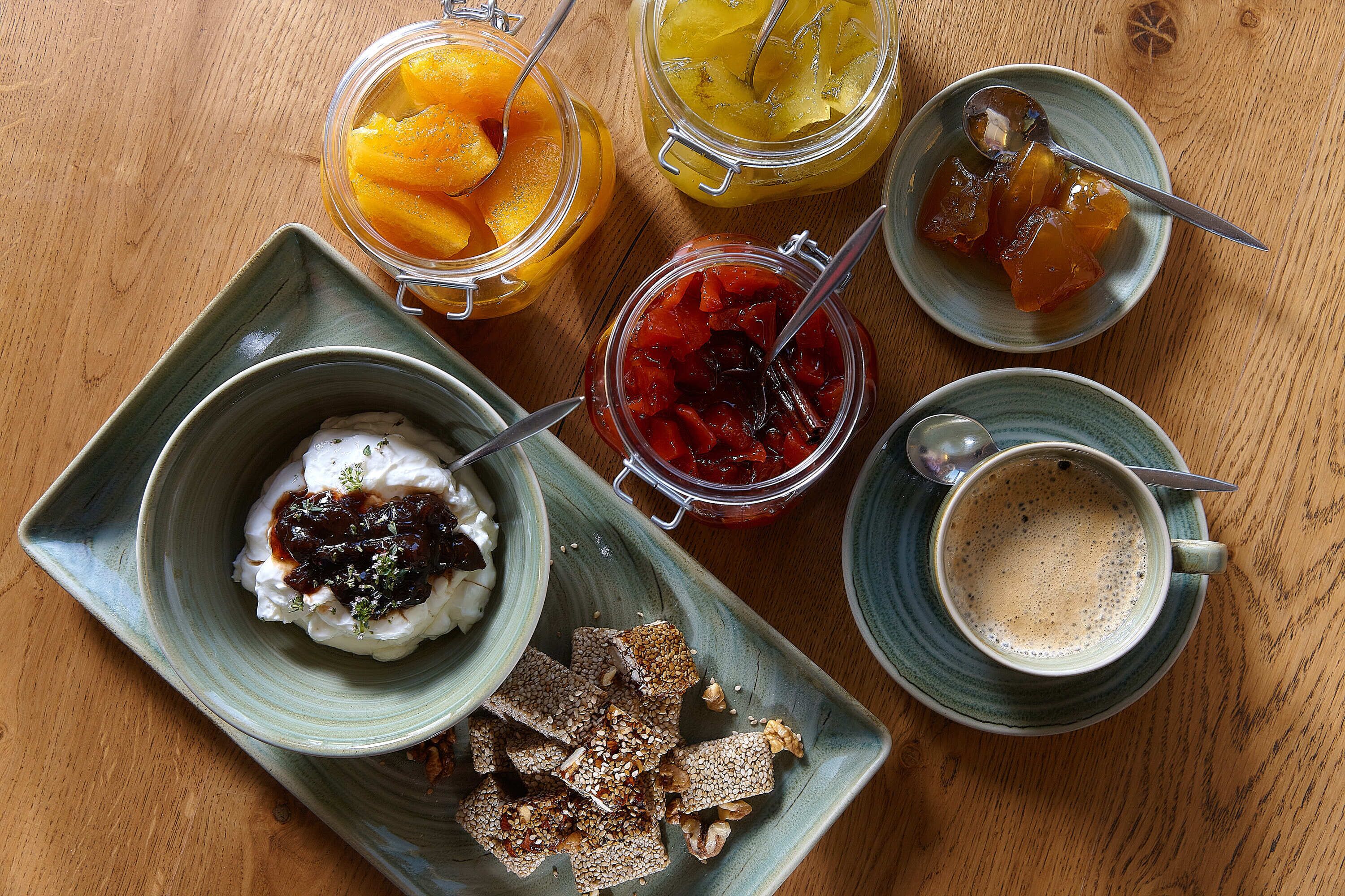 A wooden table with various food items including jars of fruit preserves, a bowl of yogurt with berries, and a cup of coffee.