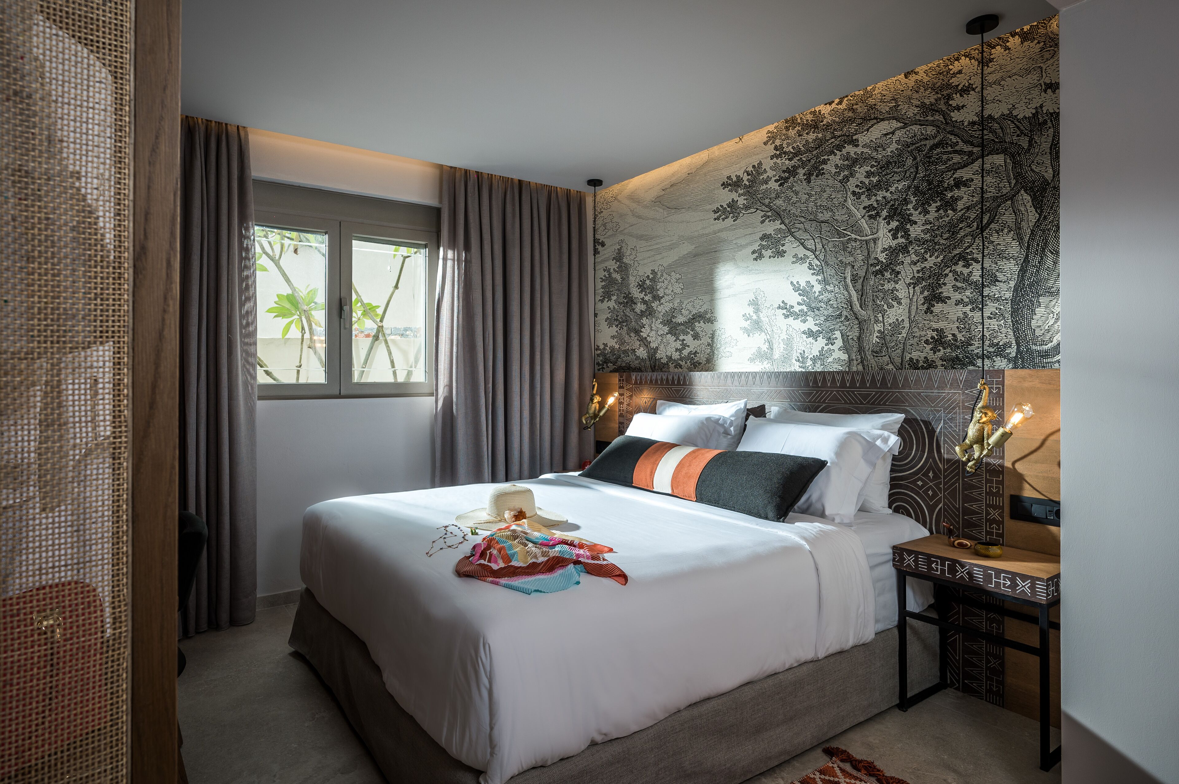 A hotel room with a large bed, a headboard with a tree mural, a nightstand with a lamp, and a window with curtains.