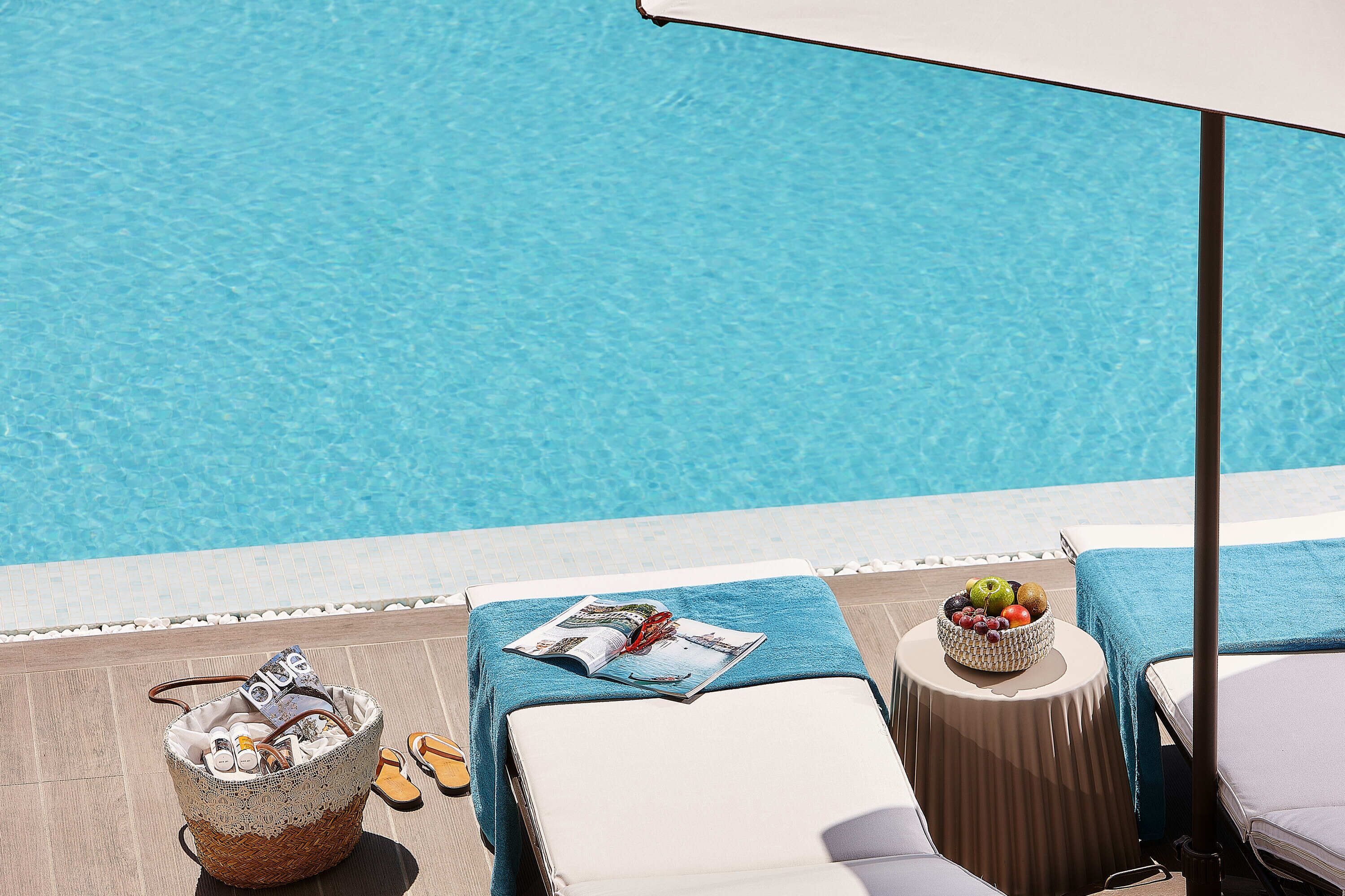 Zona junto a la piscina con una cesta de mimbre, una revista y un frutero sobre una mesita.